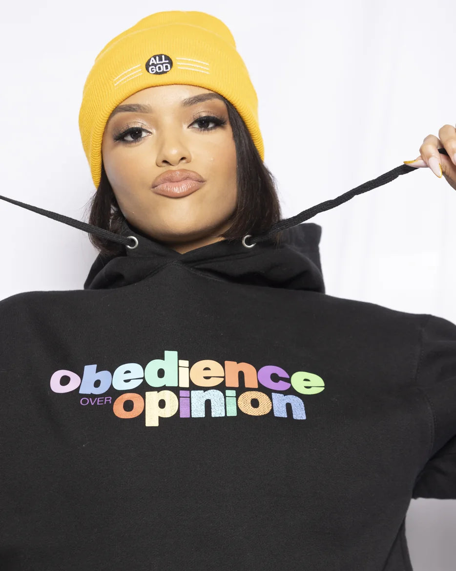 Model in a black Obedience Over Opinion hoodie and yellow All God beanie, pulling the drawstrings.